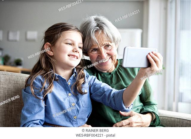 Grandmother and her granddaughter sitting together on the couch taking selfie with smartphone