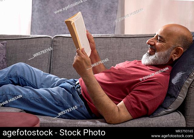 Senior man reading a book while lying on sofa at home
