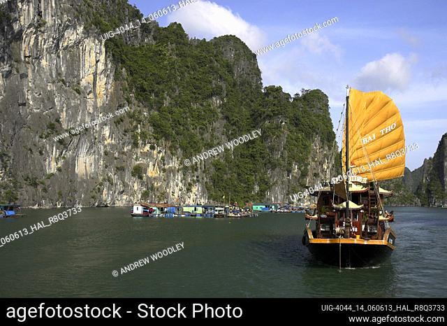 Traditional style cruising junk sails close by floating fishermens village beneath rocky islands of Halong Bay Vietnam