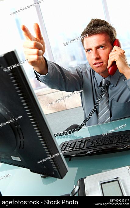 Serious businessman talking on landline phone gesturing out of picture sitting at office desk