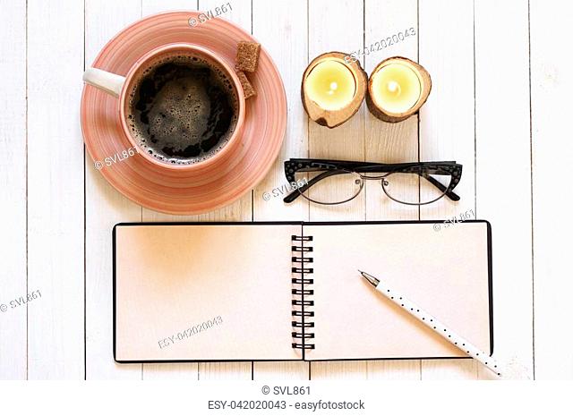 Blank notepad with pen, cup of coffee, glasses and candles on white desk. Top view point, flat lay