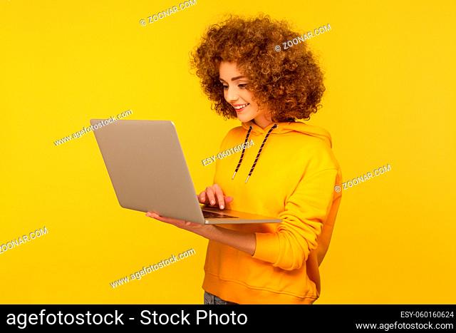 Optimistic bright woman with fluffy curly hair in urban style hoody typing on keyboard and browsing internet, working on laptop doing freelance job
