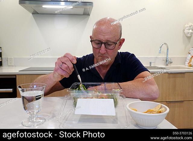 A man eats a take-out salad alone in a kitchen