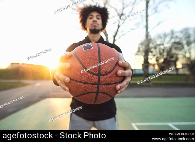 Young sportive man posing holding basketball on court at sunset