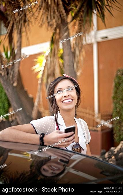 Smiling woman with mobile phone looking away while standing by car
