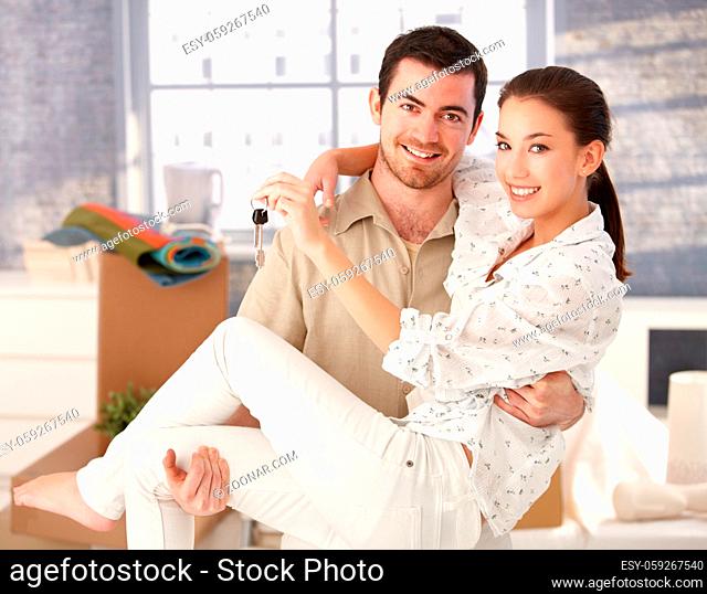 Young couple smiling happily in new house, holding keys in hand, mess around