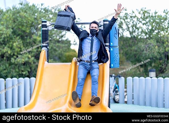 Carefree businessman wearing face mask playing on slide at park during Covid-19