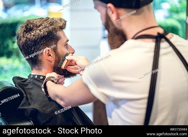 Close-up of the hands of a skilled barber using a handless brush with boar bristles, while grooming the beard of his young customer in a trendy hair salon for...