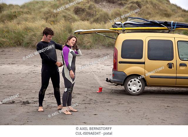 Man helping woman put on wet suit on beach