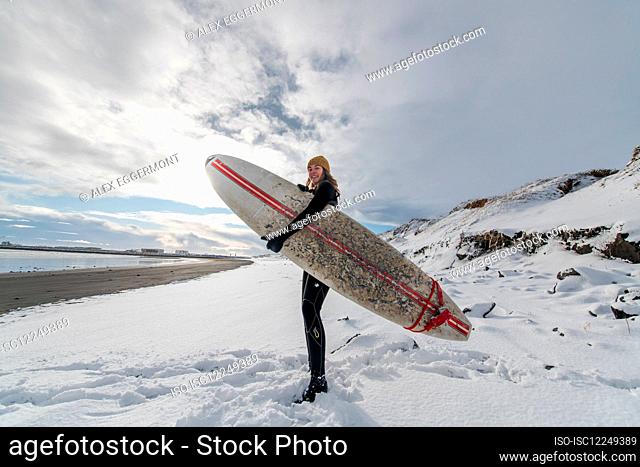 A woman wearing a wetsuit and holding a surfboard standing on a snowy beach and looking out to sea