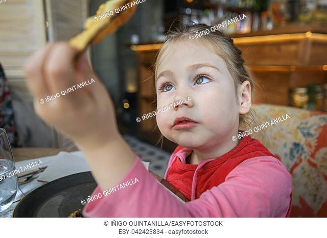 portrait of four years old blonde girl eating and looking at a fried eggplant slice in her hand, sitting in restaurant