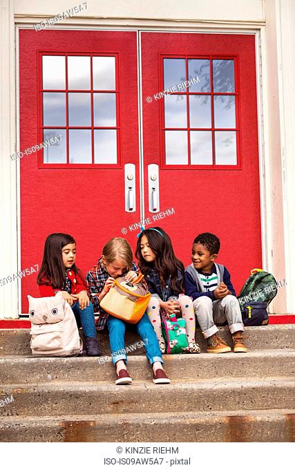 Elementary schoolgirls and boys sitting on elementary school doorway stairs looking in satchel