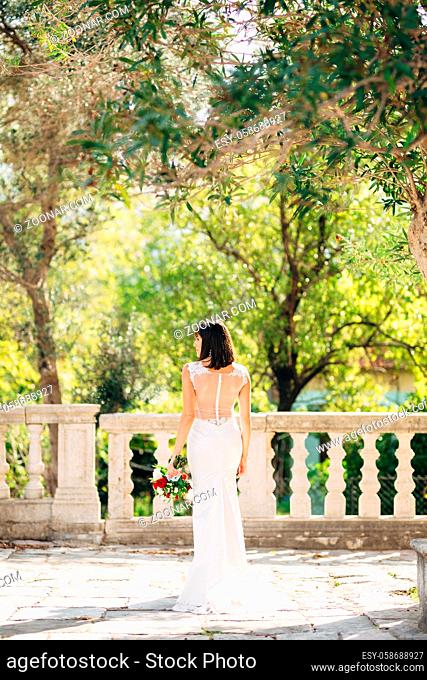 Bride in a white dress with a bouquet of flowers stands with her back on the balcony against the background of green trees. High quality photo