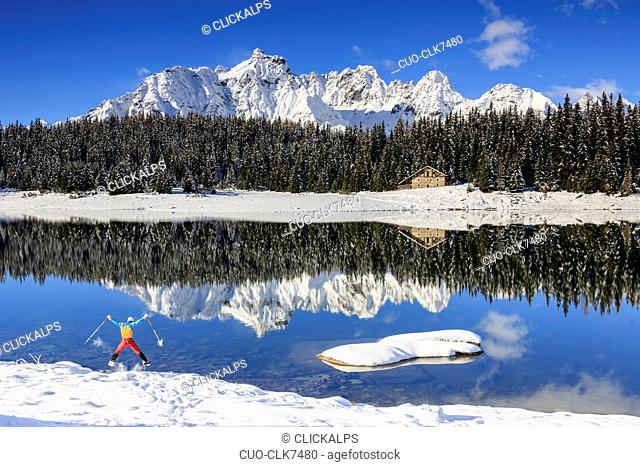 Hiker jumps on the shore of Palù Lake surrounded by snowy peaks and woods, Malenco Valley landscape, Valtellina, Lombardy, Italy, Europe