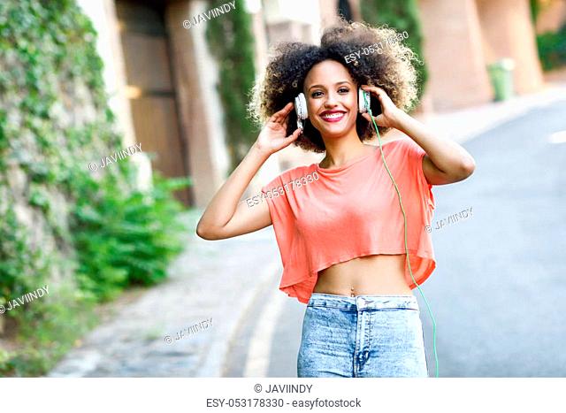 Portrait of young attractive black girl in urban background listening to the music with headphones. Woman wearing orange t-shirt and blue jeans with afro...