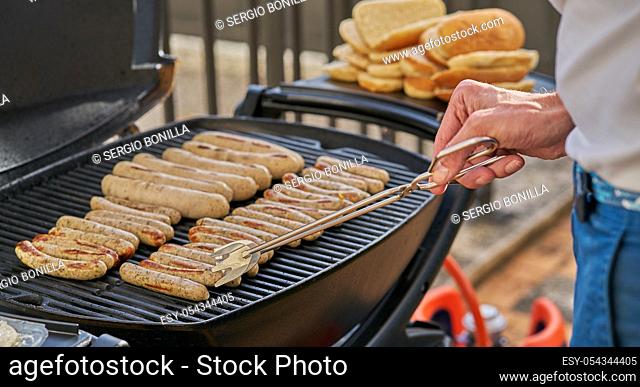 Grilled Sausage on the flaming Grill. BBQ. Bearbeque outdoors. Lots of sausages on the barbecue and you can see the chef's hand with the pincer prepared to...