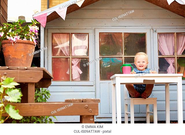 Baby girl sitting at table in front of playhouse looking at camera smiling