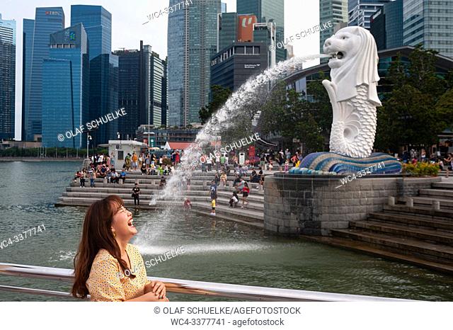 Singapore, Republic of Singapore, Asia - A female tourist poses for photos in the Merlion Park along the Singapore River with the Central Business District in...