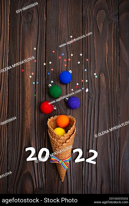 Flat lay view at Waffer cones with rainbow color balls, 2022 digits and confetti on wooden background. Lgbtq party concept