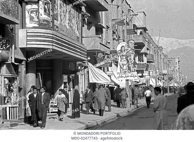 Some passers-by walking in a busy street of the city with a cinema. Outside the cinema, the poster of the film The Miller's Beautiful Wife