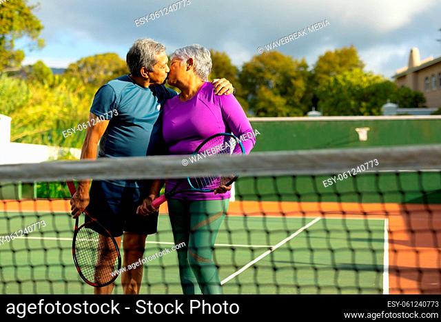 Biracial loving senior couple holding rackets kissing while standing in tennis court against sky
