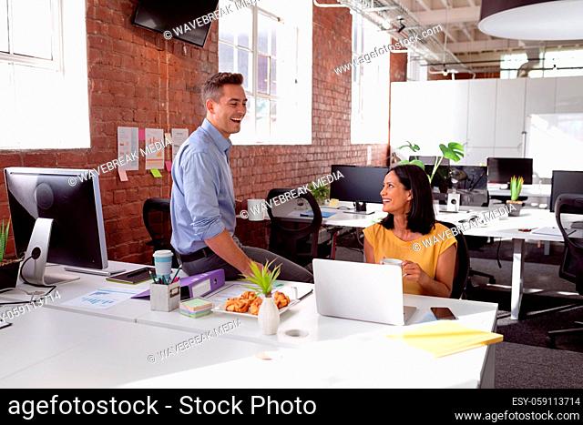 Caucasian male and female colleague sitting in office having coffee, talking and laughing