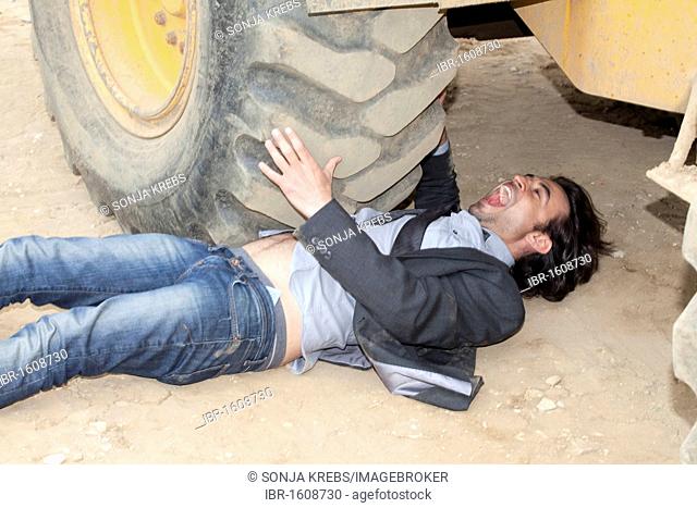 Young man lying under the wheel of an excavator, trying to free himself