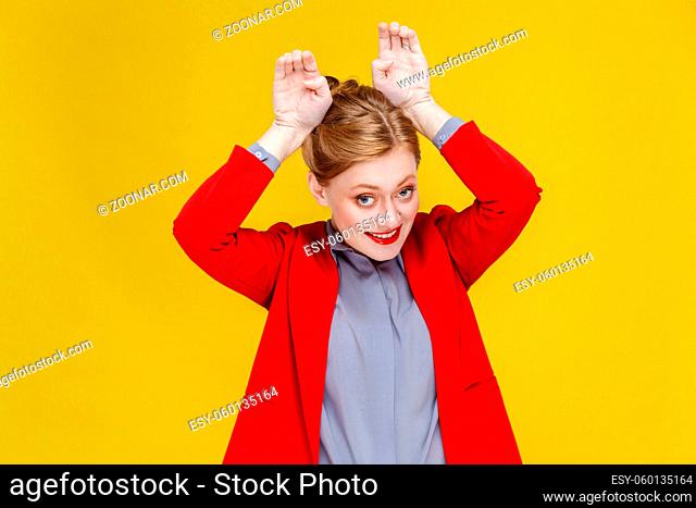 Funny business woman in red suit showing rabbit sign. Studio shot, isolated on yellow background