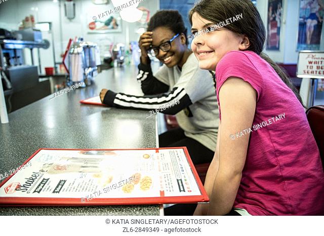 Pre-teen and teen in a diner laughing and reading a menu, eating, having a fun girls times, drinking a milkshake and playing music on the jukebox