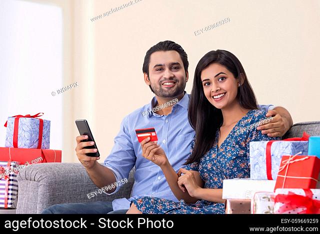 A young man and woman making online payment with credit card and smartphone in their room with gifts beside