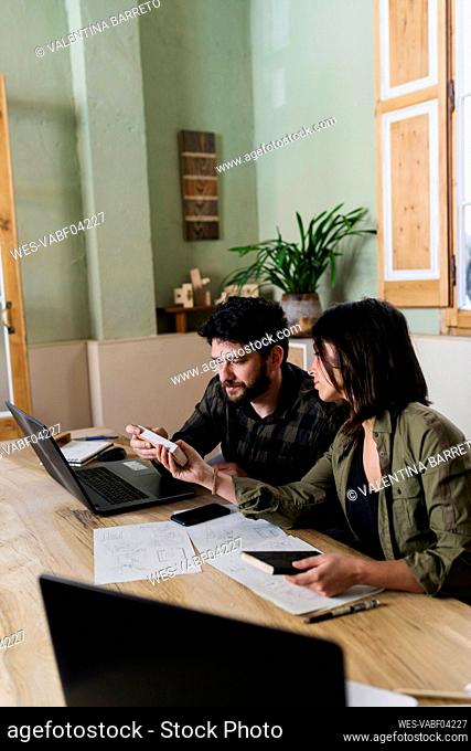 Female carpenter showing wooden samples to colleague sitting at desk in workshop