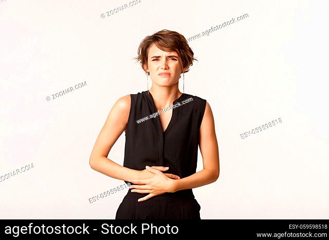 Woman in formal outfit touching belly and grimacing from pain. Girl having painful cramps or stomach ache, standing white background feeling discomfort