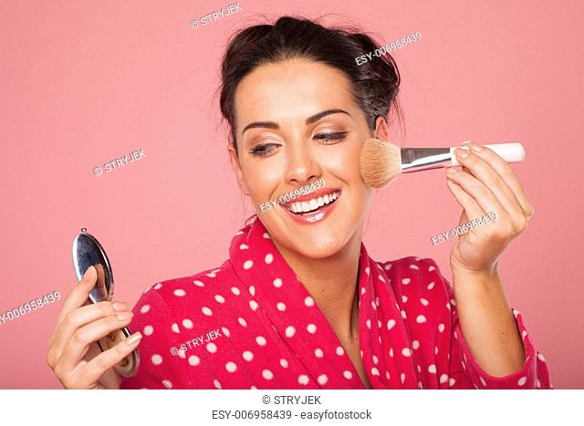 Beautiful laughing young woman applying blusher to her cheek with a large soft cosmetics brush using a handheld mirror