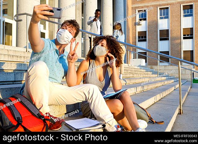 Male friend gesturing peace while taking selfie with university student in protective mask on staircase in city