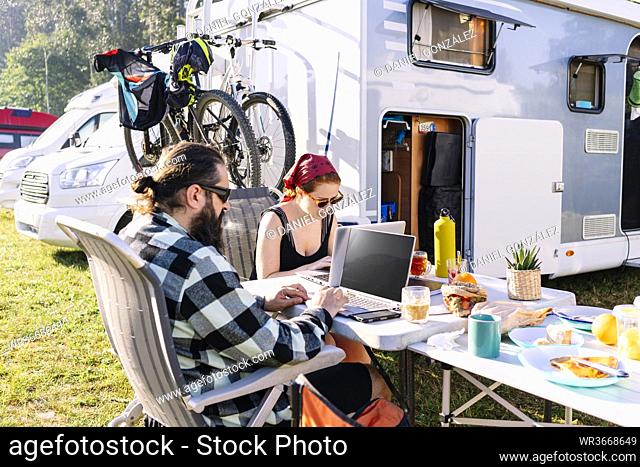 Couple sitting next to camper using laptops on table