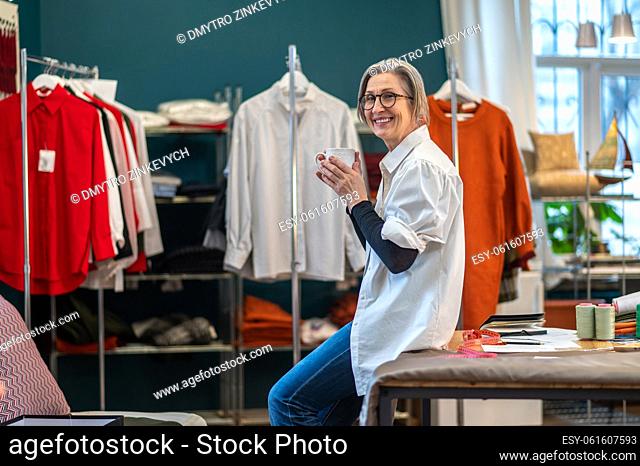 Tea time. Pretty gray-haired woman in glasses with cup crouched on table smiling at camera in sewing workshop