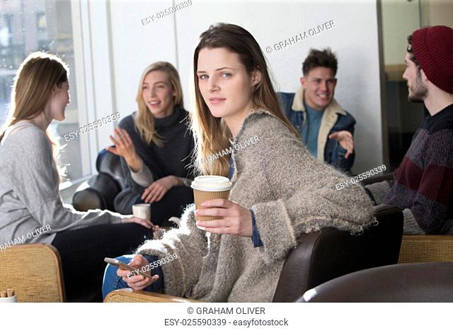 Young woman is smiling for the camera in a cafe with her friends in the background. She is holding a smartphone and a cup of coffee