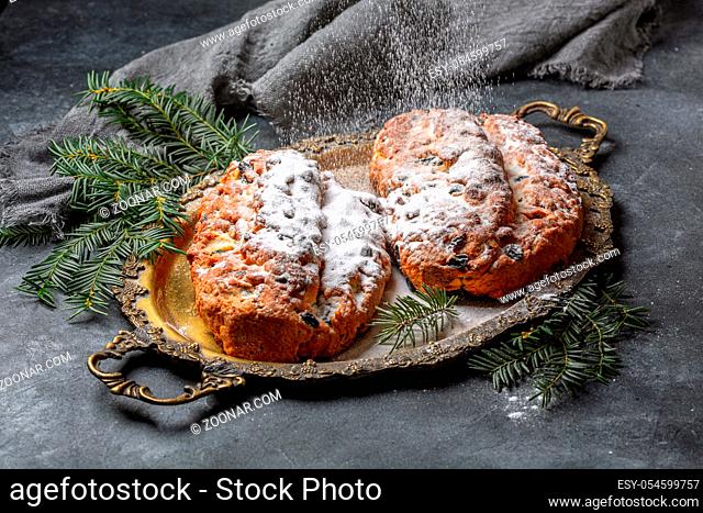 Sprinkled with powdered sugar on a homemade German Christmas Stollen in a metal tray and green fir branches on a textured dark background, selective focus