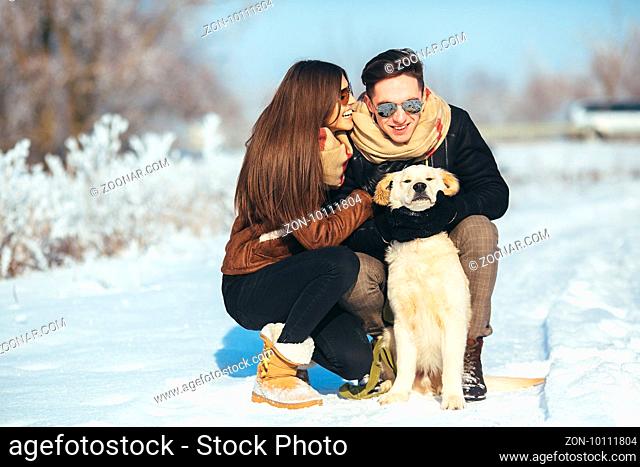 Cute young hipster couple having fun in winter park with their dog on a bright day and smiling