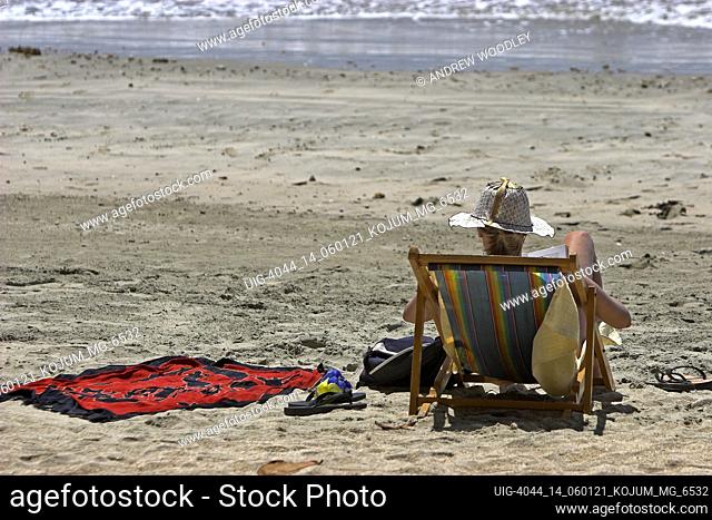 Woman in local handicraft sun hat reads book in deck chair on beach Ko Jum island Thailand