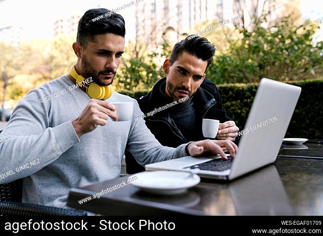 Male friends looking at laptop while having coffee sitting at sidewalk cafe at street