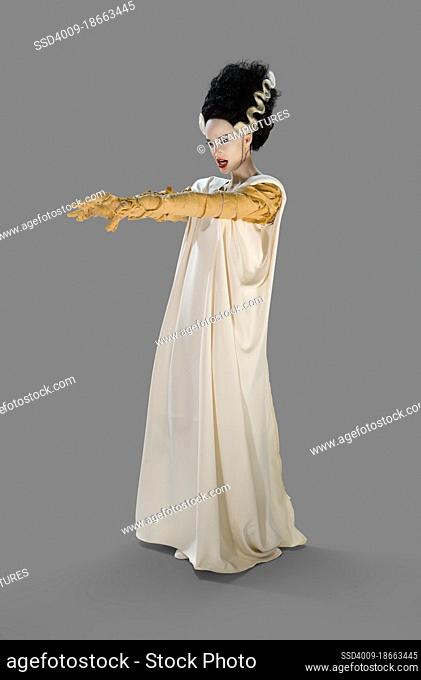 Woman dressed as the Bride Of Frankenstein Halloween costume, holding her arms out looking off camera, against a gray background