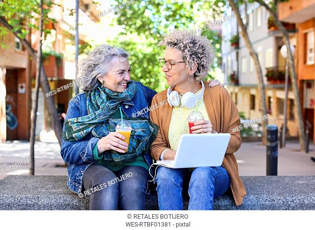Senior mother with her adult daughter using laptop and drinking juices in the city