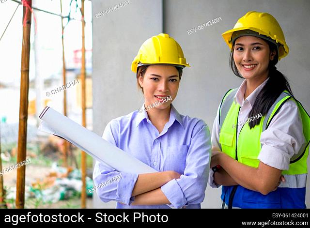 Young woman in construction helmet and floor plan stand with arms crossed at the construction site of housing projects. Asian engineer in a safety vest stand...