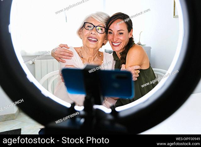 Smiling granddaughter and grandmother sitting with arm around while vlogging through smart phone at home