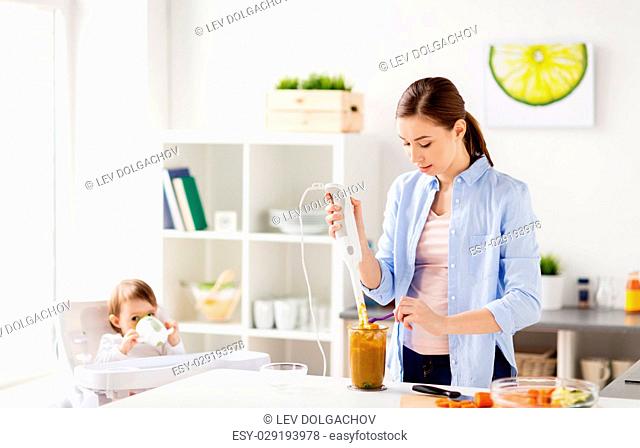 family, food, child, eating and parenthood concept - mother with blender cooking puree for her baby sitting in highchair at home kitchen
