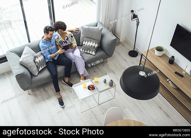 Multi-ethnic couple using digital tablet while sitting on sofa in living room of modern penthouse