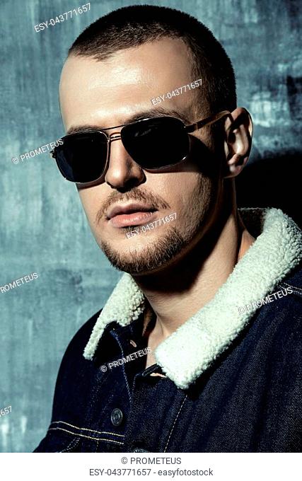 Sexual male model posing in jeans clothes and sunglasses. Short hair styling. Studio fashion shot