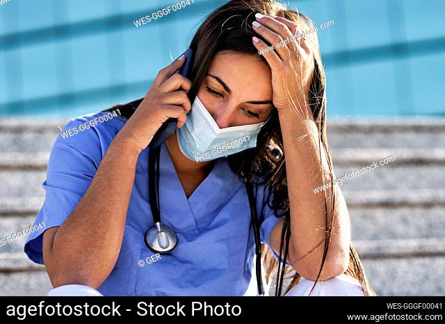 Young doctor wearing face mask talking on mobile phone while sitting on staircase against hospital