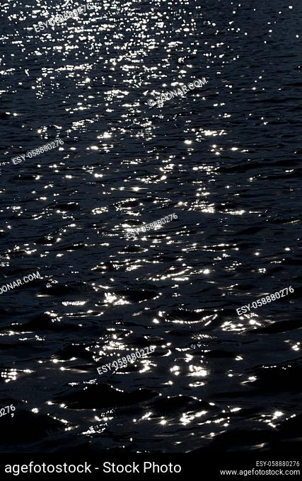 black abstract background of dark water in a lake and white sun glare on the surface of waves, out of focus